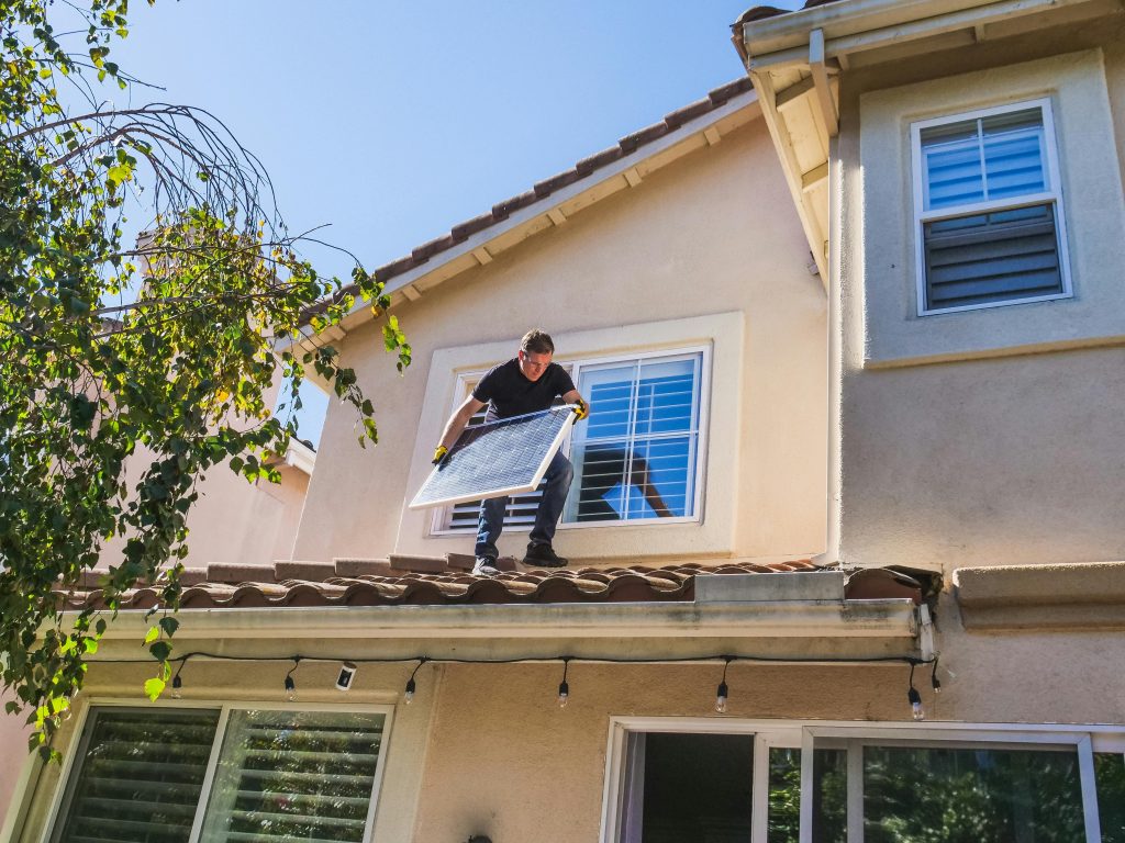 A person in a black shirt and jeans, wearing yellow gloves, is standing on a terracotta tile roof and carefully holding a square solar panel. They are in the process of installing it on the upper-story window of a two-story stucco house.